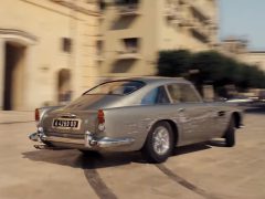 Een zilveren Aston Martin DB9 die door een stadsstraat rijdt, te zien in No Time To Die.