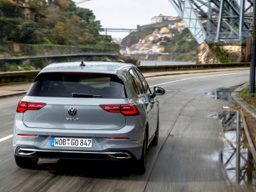 De Volkswagen Golf 8 rijdt over een weg met een brug op de achtergrond.
