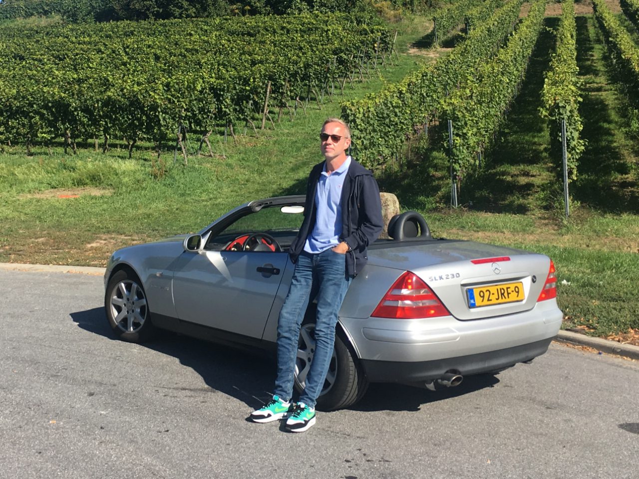 Een man die naast een zilveren auto staat voor een wijngaard op AutoRAI.nl.