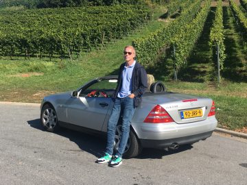 Een man die naast een zilveren auto staat voor een wijngaard op AutoRAI.nl.