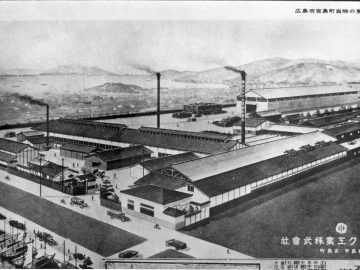 Industriële faciliteiten met Mazda-schoorstenen die rook uitstoten, tegen de achtergrond van een bergachtig landschap, op een historische zwart-witfoto.