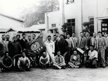 Groep mannen uit het begin van de 20e eeuw poseert met een Mazda-motorfiets en vlaggen voor een gebouw.