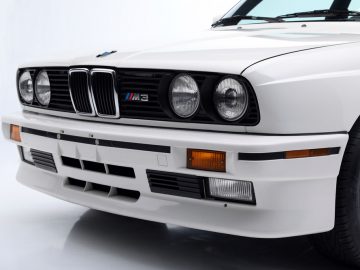 Hoekaanzicht van voren van een witte BMW M3 (E30) met de grille en koplampen, die doet denken aan de serie "The Fast and the Furious".