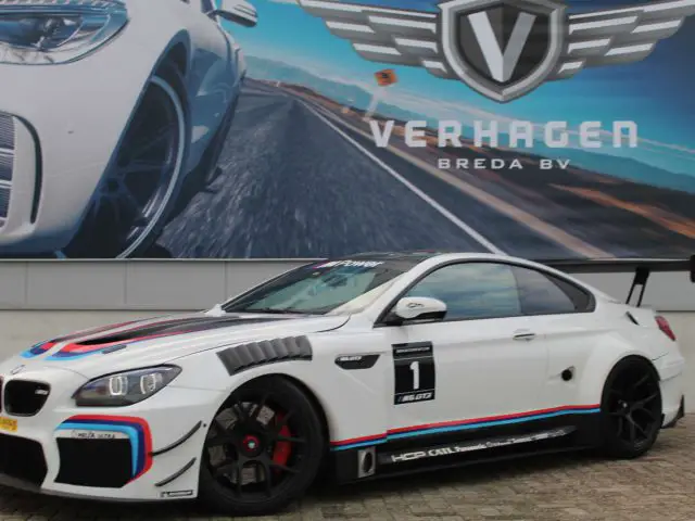 Witte BMW M6 GT3-racewagen met sponsorstickers geparkeerd voor een reclamebord.