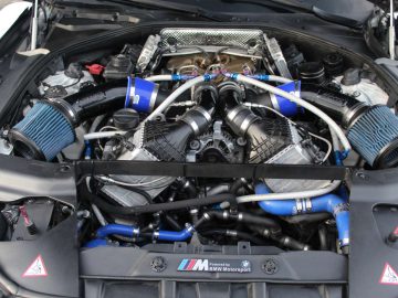 Een open motorkap onthult een gemodificeerde BMW M6 GT3-automotor met aftermarket-onderdelen en blauwe siliconenslangen.