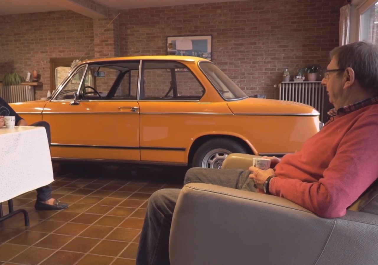 Twee individuen zitten en praten met een vintage BMW die in de woonkamer geparkeerd staat.