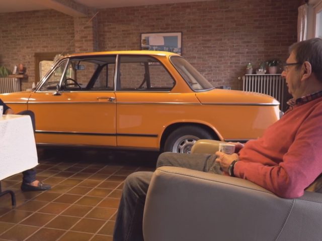 Twee individuen zitten en praten met een vintage BMW die in de woonkamer geparkeerd staat.
