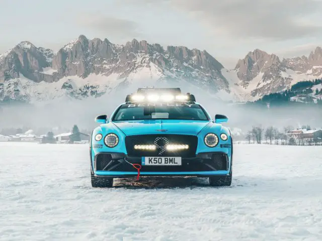 Een blauwe Bentley Continental Ice GT uitgerust met een dakdrager, geparkeerd op sneeuw, met bergachtig terrein op de achtergrond.