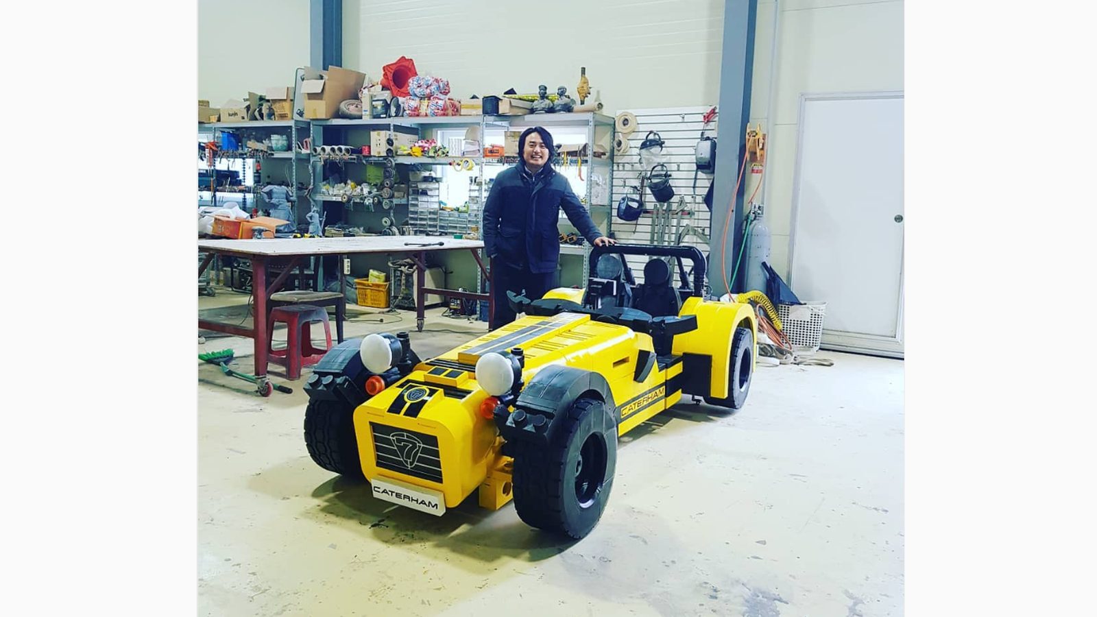 Een persoon die naast een gele Caterham 620R staat in een werkplaatsomgeving.