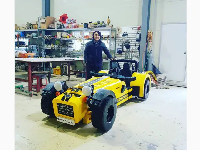 Een persoon die naast een gele Caterham 620R staat in een werkplaatsomgeving.