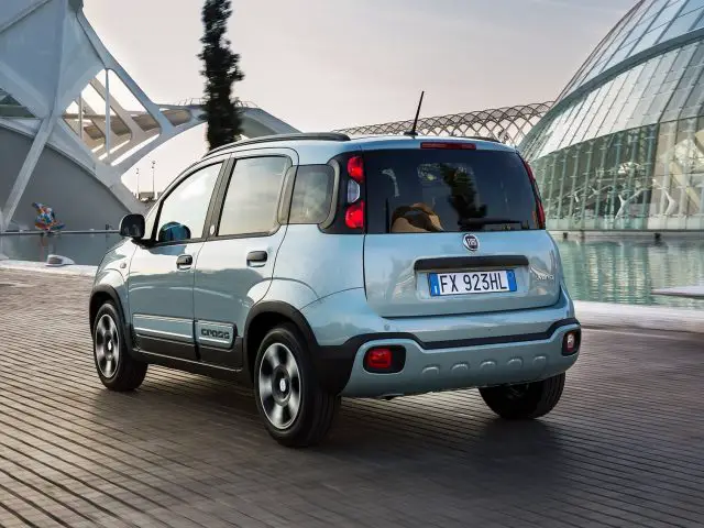 Een lichtblauwe Fiat 500 hybride auto die op een verhard terrein rijdt in de buurt van moderne architectuur.