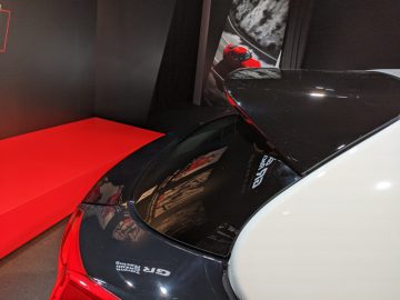 Achteraanzicht van een witte GR Yaris-sportwagen met open spoiler, tentoongesteld op een binnententoonstelling met rode accenten op de achtergrond.