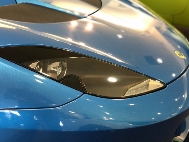 Close-up van de koplamp en het motorkapembleem van een blauwe Lotus-sportwagen.