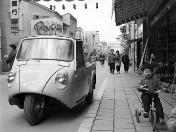 Vintage Mazda driewielige auto en kind op trapauto in een drukke straat.
