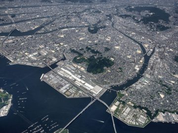 Luchtfoto van een dicht stedelijk gebied met bruggen over een rivier, industriële zones aan de waterkant en een prominente Mazda-dealer die duidelijk zichtbaar is in het tafereel.