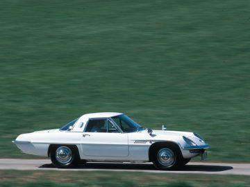 Witte vintage Mazda-sportwagen in beweging op een weg met een met gras begroeide achtergrond.