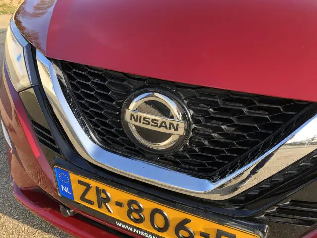 Close-up van de grille van een rode Nissan Qashqai met het embleem en een Nederlands kenteken zichtbaar.