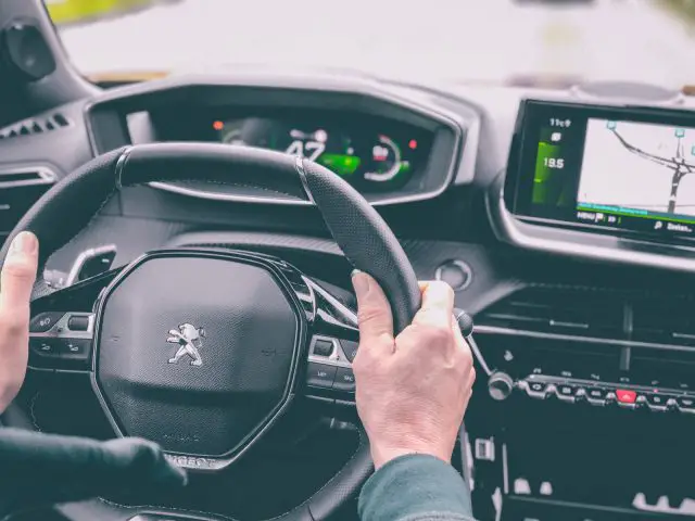 Handen van de bestuurder op het stuur van een Peugeot 2008 met zicht op het dashboard en het navigatiesysteem van de auto.