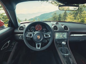 Binnenaanzicht van een luxe Porsche 718 GTS-auto met stuur en dashboard, met uitzicht op een bosrijk berglandschap.
