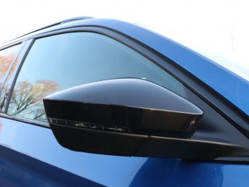 Zijaanzicht van de zijspiegel van een blauwe Skoda Superb met bomen weerspiegeld in de ramen.