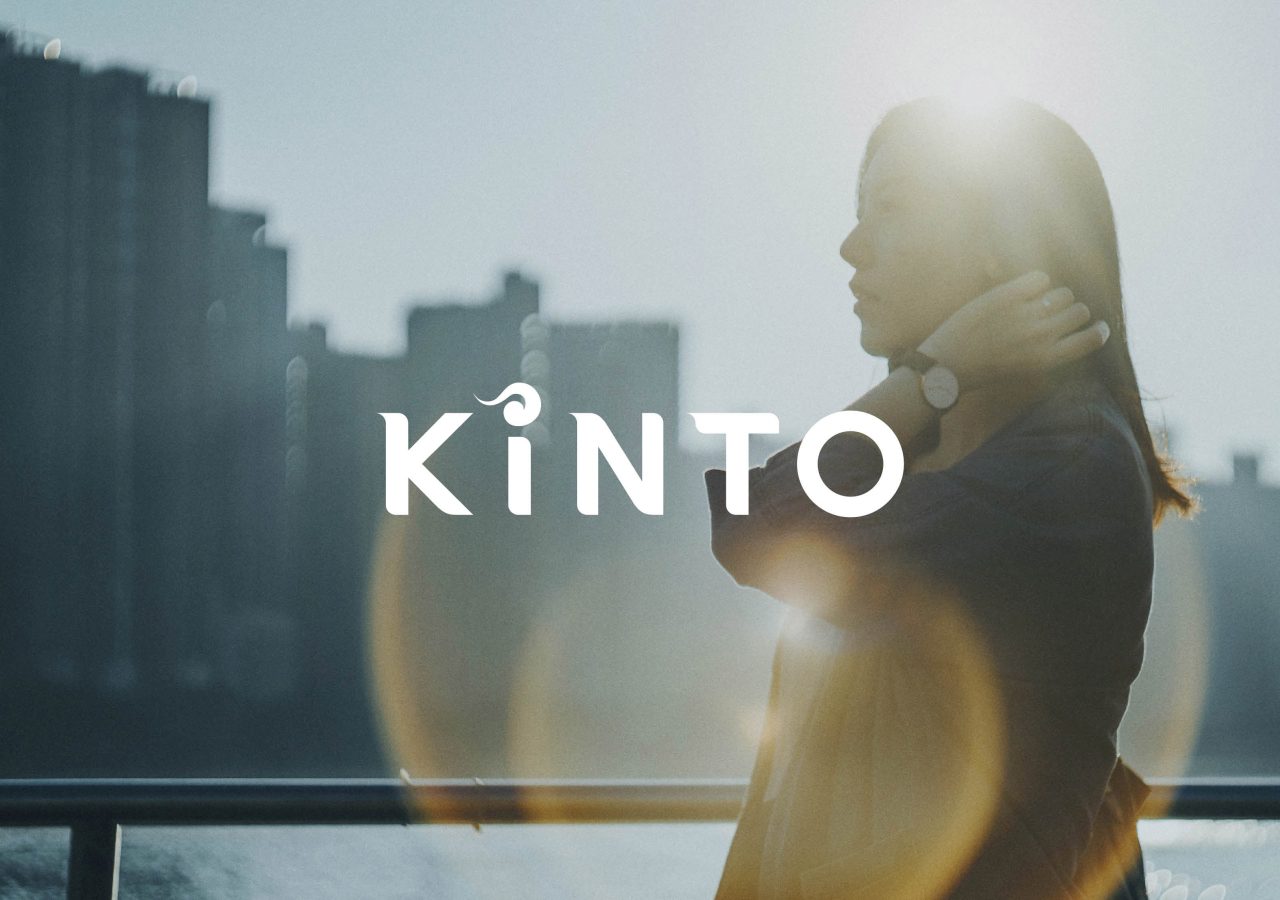 Vrouw staart in de verte met een Toyota KINTO en de skyline van de stad erachter, lensflare-effect zichtbaar.