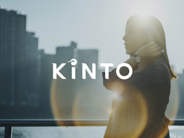 Vrouw staart in de verte met een Toyota KINTO en de skyline van de stad erachter, lensflare-effect zichtbaar.
