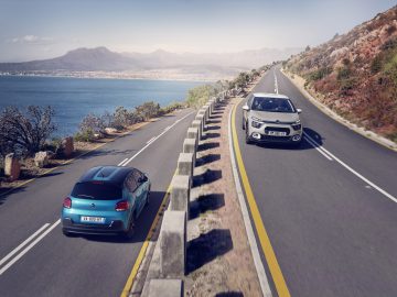 Twee Citroën C3-auto's rijden op een kustweg met een schilderachtige berg- en oceaanachtergrond.
