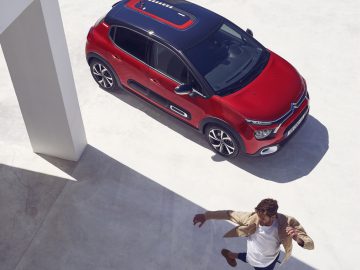 Een persoon die op een zonnige dag naar een rode Citroën C3 loopt, geparkeerd onder een schaduwrijk bouwwerk.