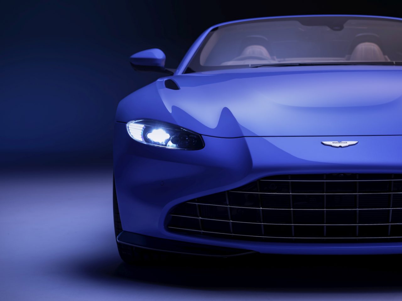 Een blauwe Aston Martin Vantage Roadster met koplampen aan, gefotografeerd in een studio met een donkerblauwe achtergrond met kleurverloop.