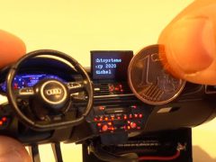 De vingers van een persoon houden een munt vast naast een zeer gedetailleerd miniatuurmodel van een Audi RS6-dashboard en stuur op schaal.