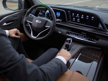 Persoon die op de bestuurdersstoel zit van een moderne Cadillac Escalade met een touchscreen-dashboard en de versnellingspook vasthoudt.