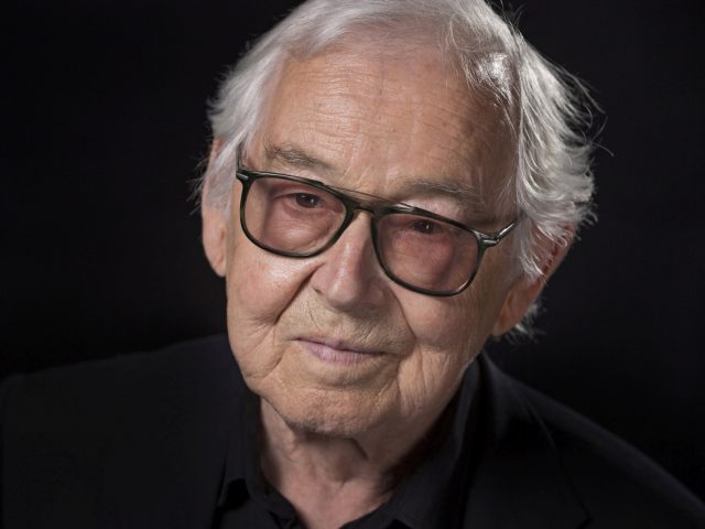 Een oudere man met een bril en wit haar, gekleed in een donker shirt, tegen een zwarte achtergrond is Erhard Schnell.