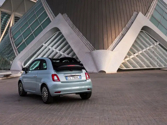 Een Fiat 500 Hybrid geparkeerd voor een modern gebouw met opvallende architectonische kenmerken.