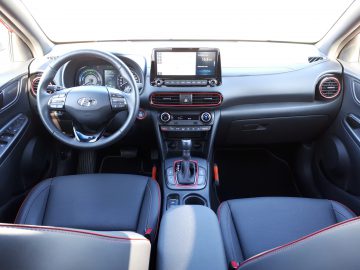 Binnenaanzicht van een moderne Kona Hybrid-auto met dashboard in cockpitstijl, digitale displays en rode accentafwerking.