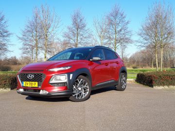 Rode Hyundai Kona Hybrid geparkeerd op een verhard terrein met bomen en groen op de achtergrond op een zonnige dag.