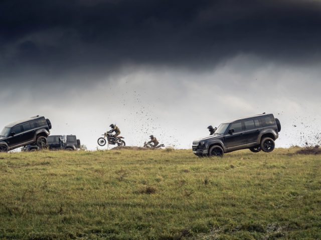 Twee Land Rover Defenders en een motorfiets voeren een offroad-stunt uit op een met gras begroeide heuvel met een stormachtige lucht op de achtergrond.