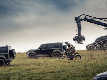 Een filmploeg legt een actiescène vast met motorrijders en Land Rover Defenders op een met gras begroeide heuvel.