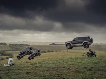 Twee Land Rover Defenders in de lucht tegen een stormachtige lucht, met een motorrijder en uitrusting op de grond beneden.