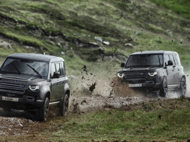 Twee Land Rover Defenders rijden door modderig terrein, opspattend water en vuil.