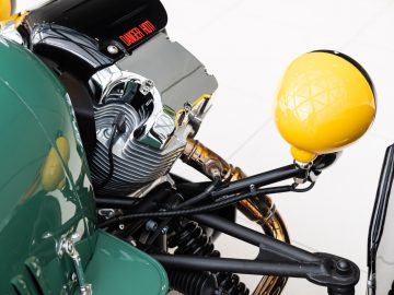 Close-up van de motor en uitlaat van een Morgan-motorfiets met een ronde, gele zijspiegel.