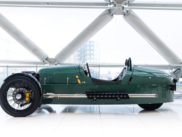 Zijaanzicht van een vintage groene Morgan-raceauto met één zitplaats, tentoongesteld in een witte moderne galerij.