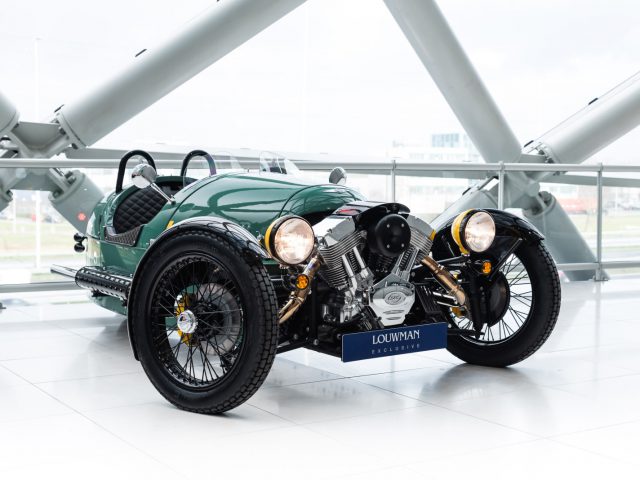 Een vintage groene Morgan-sportwagen tentoongesteld in een modern gebouw met structurele balken boven het hoofd.