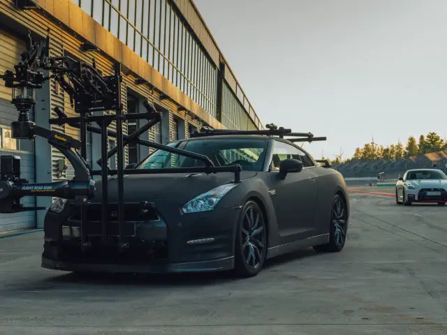 Een zwarte Nissan GT-R-sportwagen uitgerust met een professionele camera-installatie, geparkeerd op een racecircuit, met een andere sportwagen op de achtergrond.