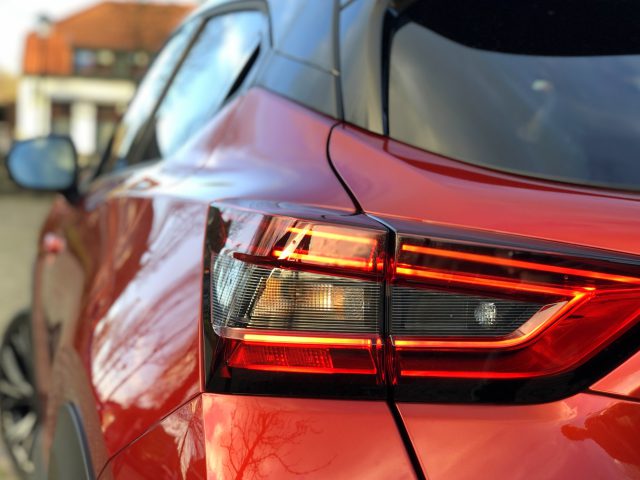 Close-up van het rode achterlicht en het achterste zijpaneel van een Nissan Juke, met een onscherpe achtergrond van een stadslandschap.