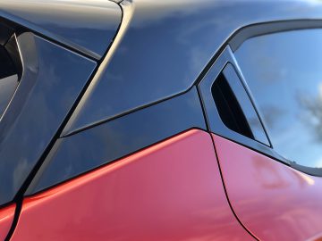 Close-up van de carrosserie van een Nissan Juke met een contrast tussen rode en zwarte panelen onder een blauwe lucht.