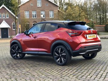 Rode Nissan Juke geparkeerd op geplaveide stoep vlakbij bakstenen gebouwen.