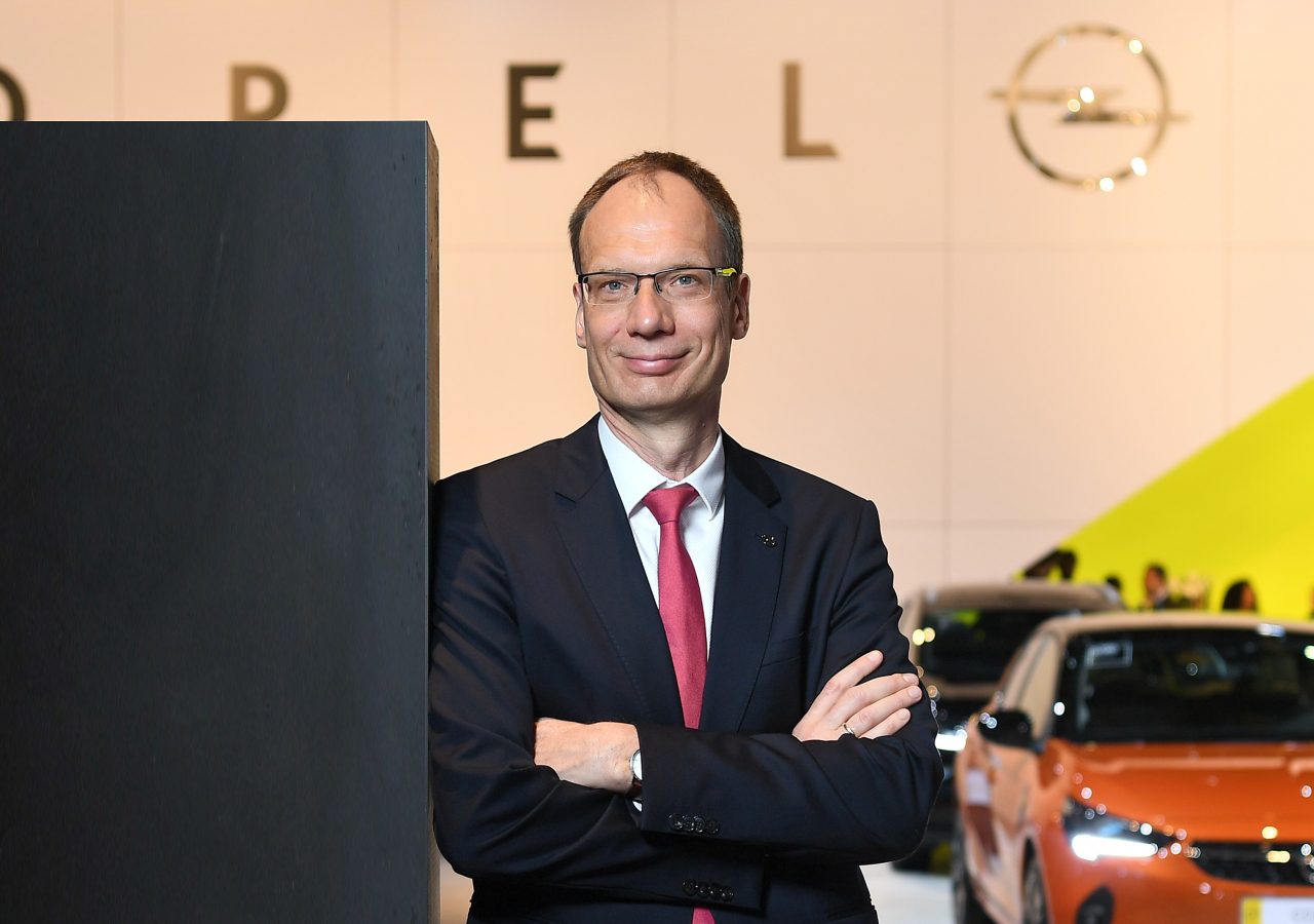 Michael Lohscheller, een man in pak met gekruiste armen, staat zelfverzekerd op een bedrijfsevenement met het logo van zijn bedrijf op de achtergrond.