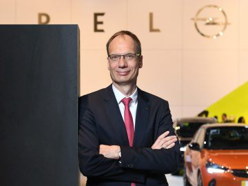 Michael Lohscheller, een man in pak met gekruiste armen, staat zelfverzekerd op een bedrijfsevenement met het logo van zijn bedrijf op de achtergrond.