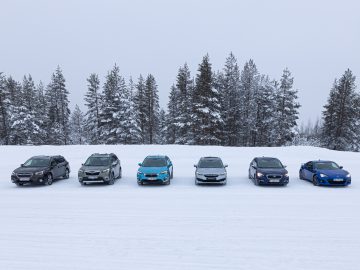 Een opstelling van verschillende auto's, waaronder een Subaru Forester, geparkeerd op een met sneeuw bedekt veld met op de achtergrond dichte groenblijvende bomen onder een bewolkte hemel.