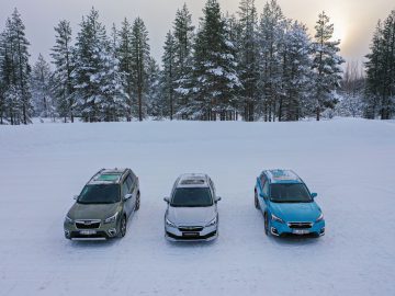 Drie Subaru Forester-auto's geparkeerd op een met sneeuw bedekte open plek met een achtergrond van pijnbomen en een bewolkte hemel.